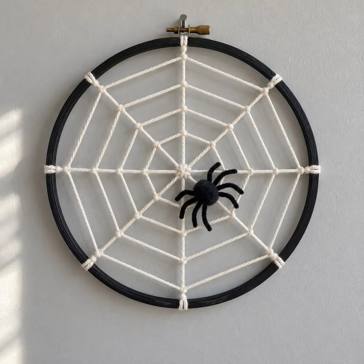 Cream yarn spiderweb with eight spokes inside a black embroidery hoop, fuzzy black spider, hung on a gray wall