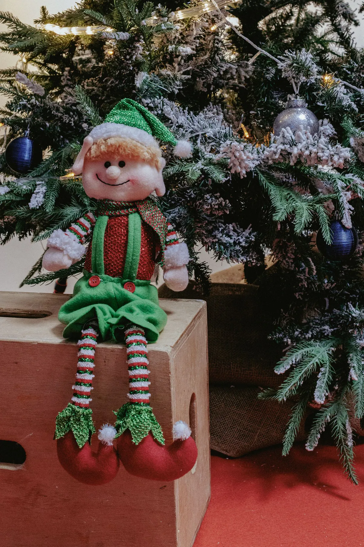 Plush elf in a green glitter hat sitting on a small wooden box beside a flocked Christmas tree