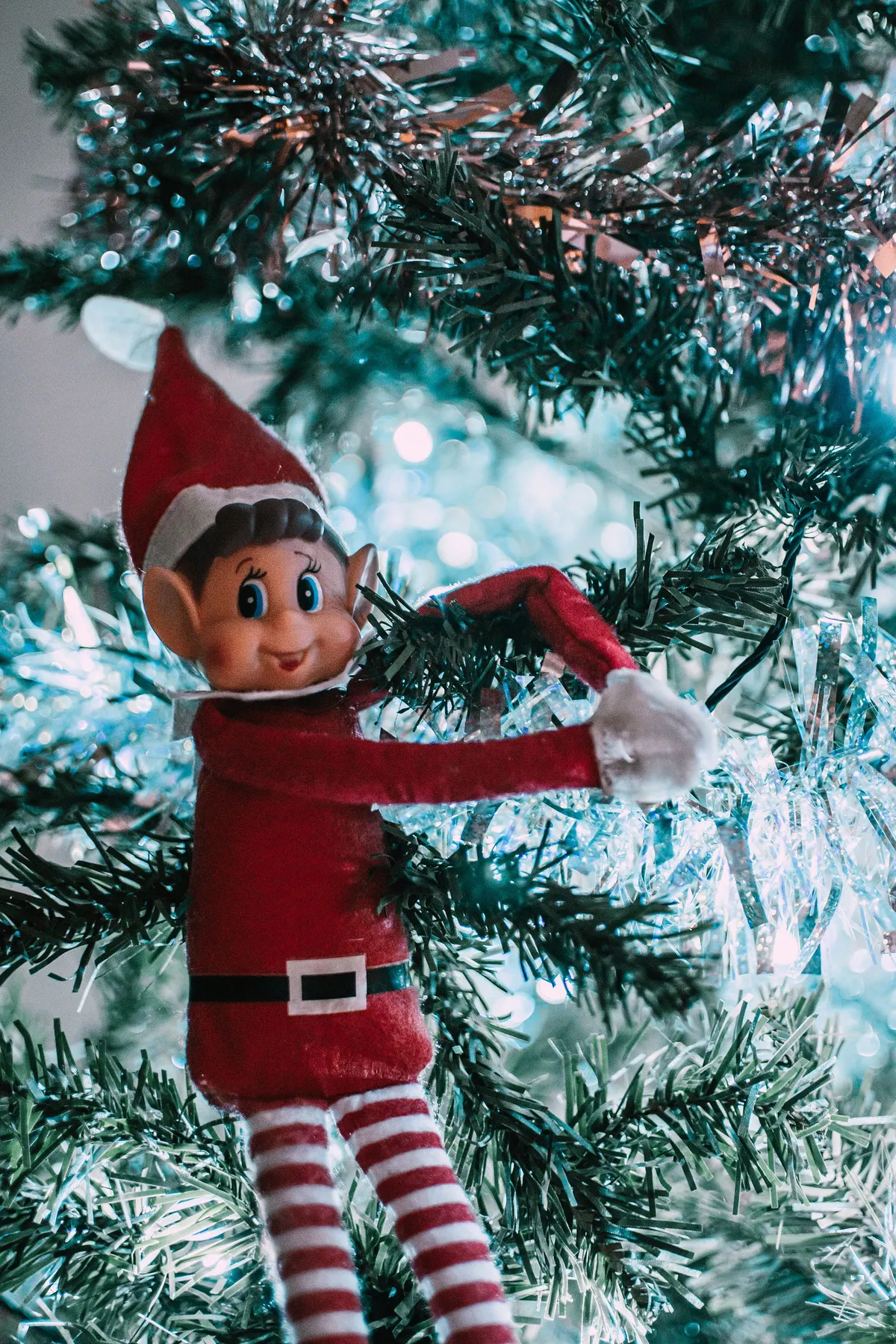 Small elf in a red suit and striped stockings hugging a tree branch among silver tinsel and lights