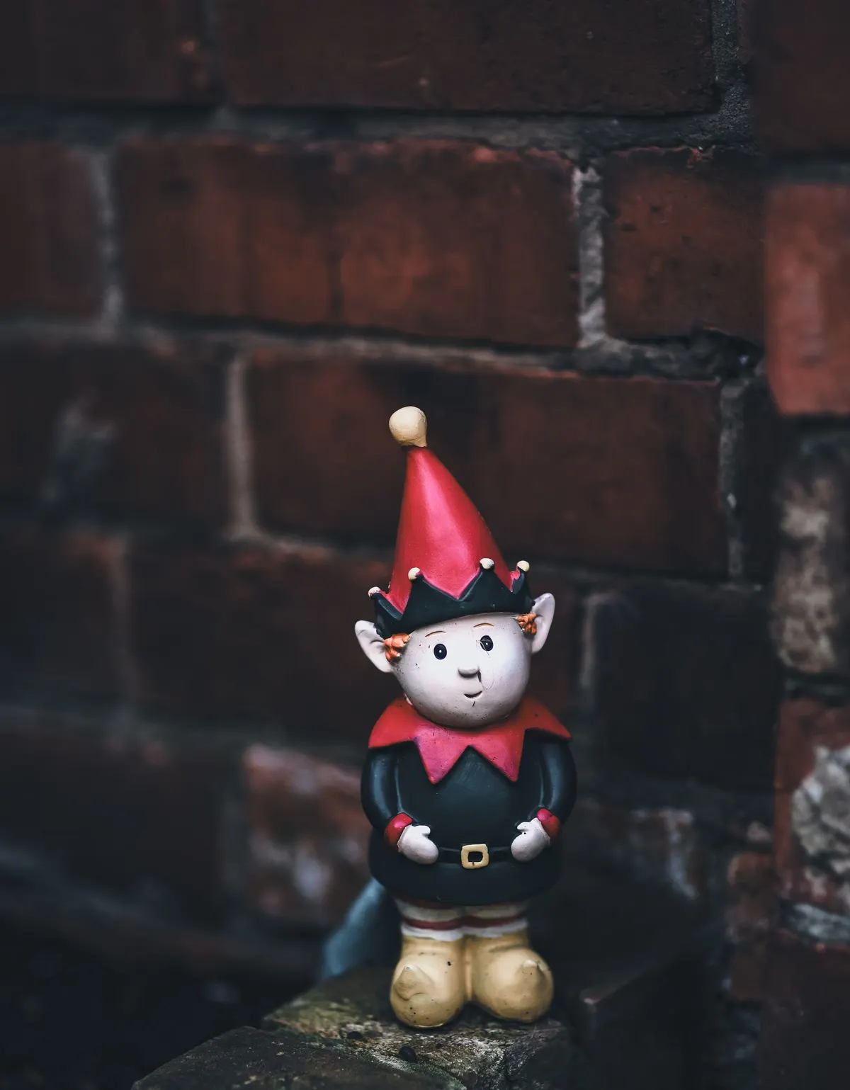 Elf figurine in a red pointed hat, green suit and yellow boots standing on a brick ledge