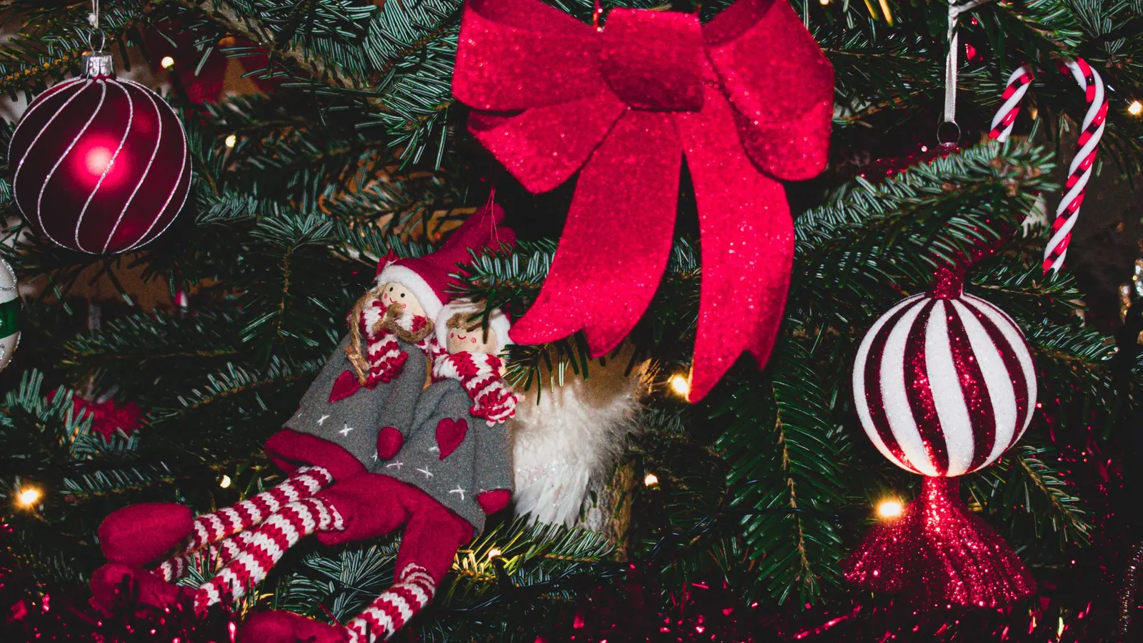 Two cloth elf dolls with red hats and striped legs lounging on Christmas tree branches under a red bow