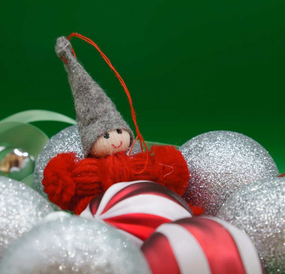 Small yarn elf ornament with a gray felt hat tucked among silver glitter and striped baubles