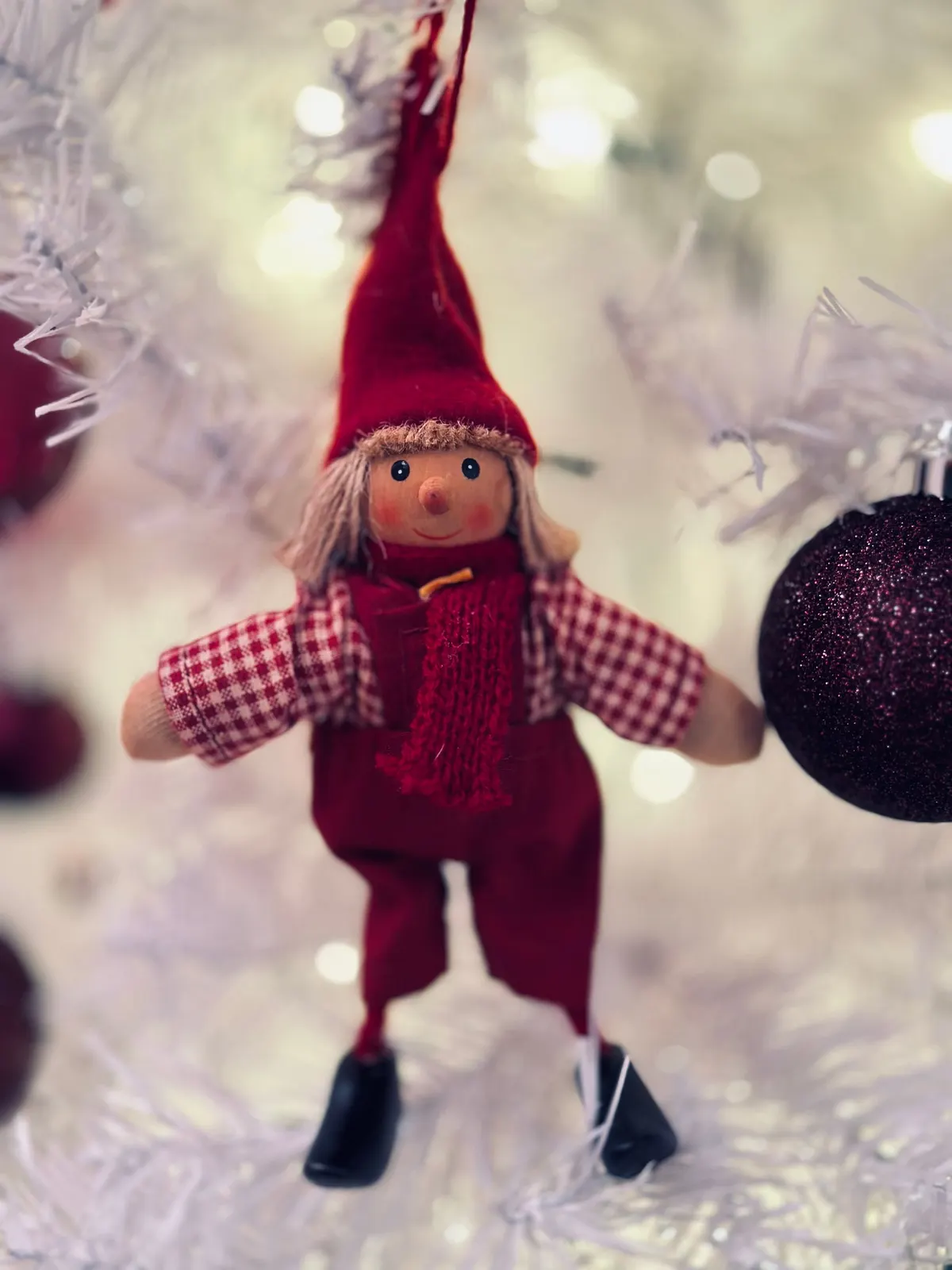 Elf ornament with a tall red hat and checked sleeves hanging beside a plum glitter bauble on a white tree