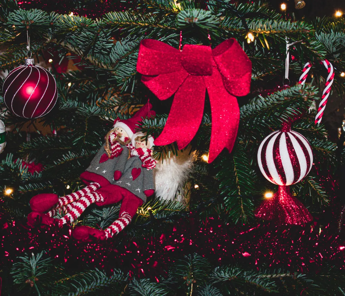 Two cloth elf dolls with red hats and striped legs lounging on Christmas tree branches under a red bow