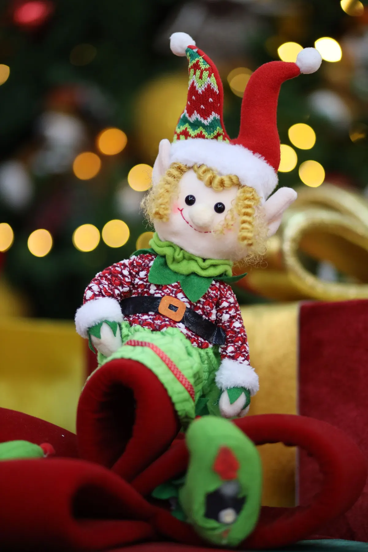 Plush elf with blond curls and curled red shoes lounging in loops of red velvet ribbon by a tree