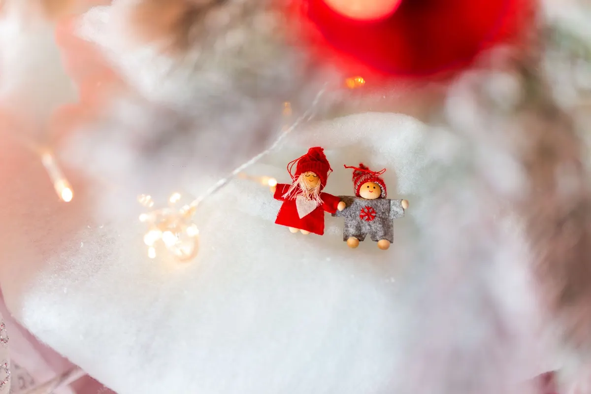 Two tiny red-hatted elf figures lying with arms out on white cotton snow beside a string of lights