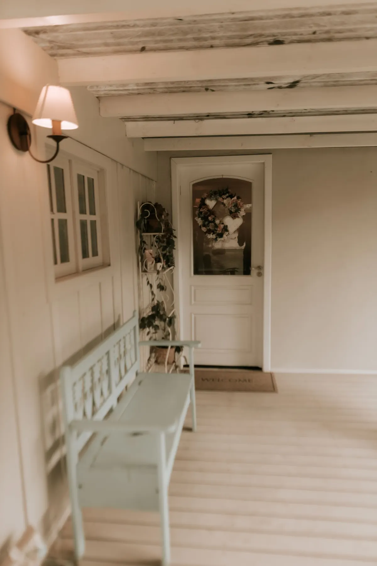 Pale wooden bench on a white covered porch beside a door with a wreath and doormat