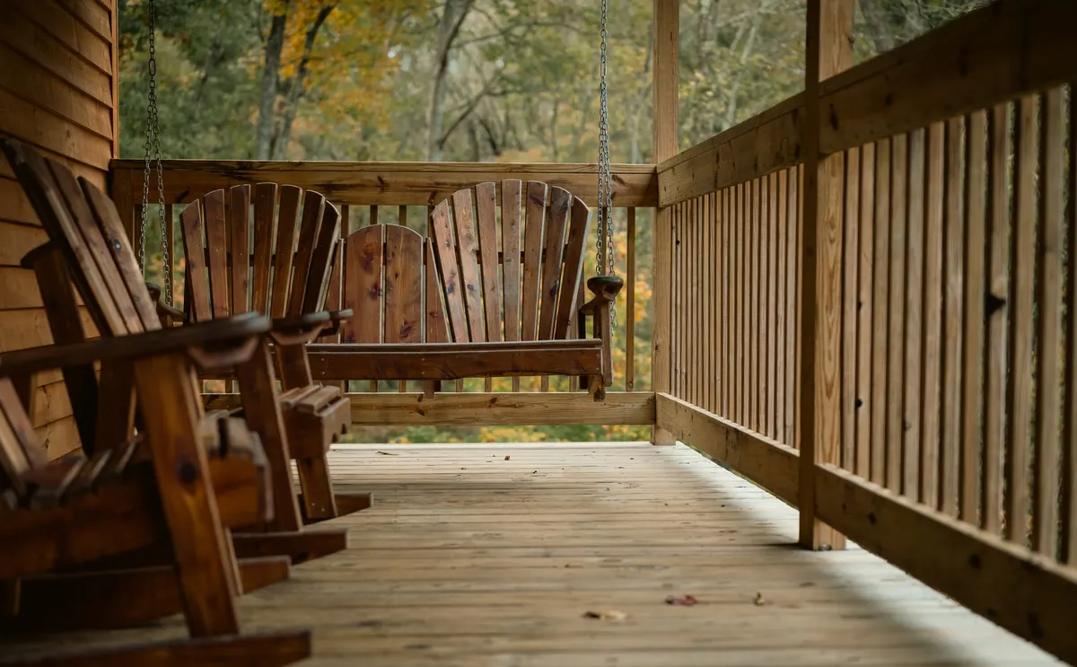 Wooden covered porch with rocking chairs and a hanging slatted swing, autumn trees beyond the railing