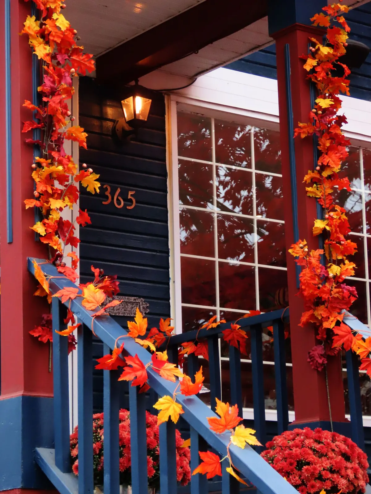 Orange leaf garlands wrapped around white porch posts and a blue railing, with red mums and a lit wall lantern