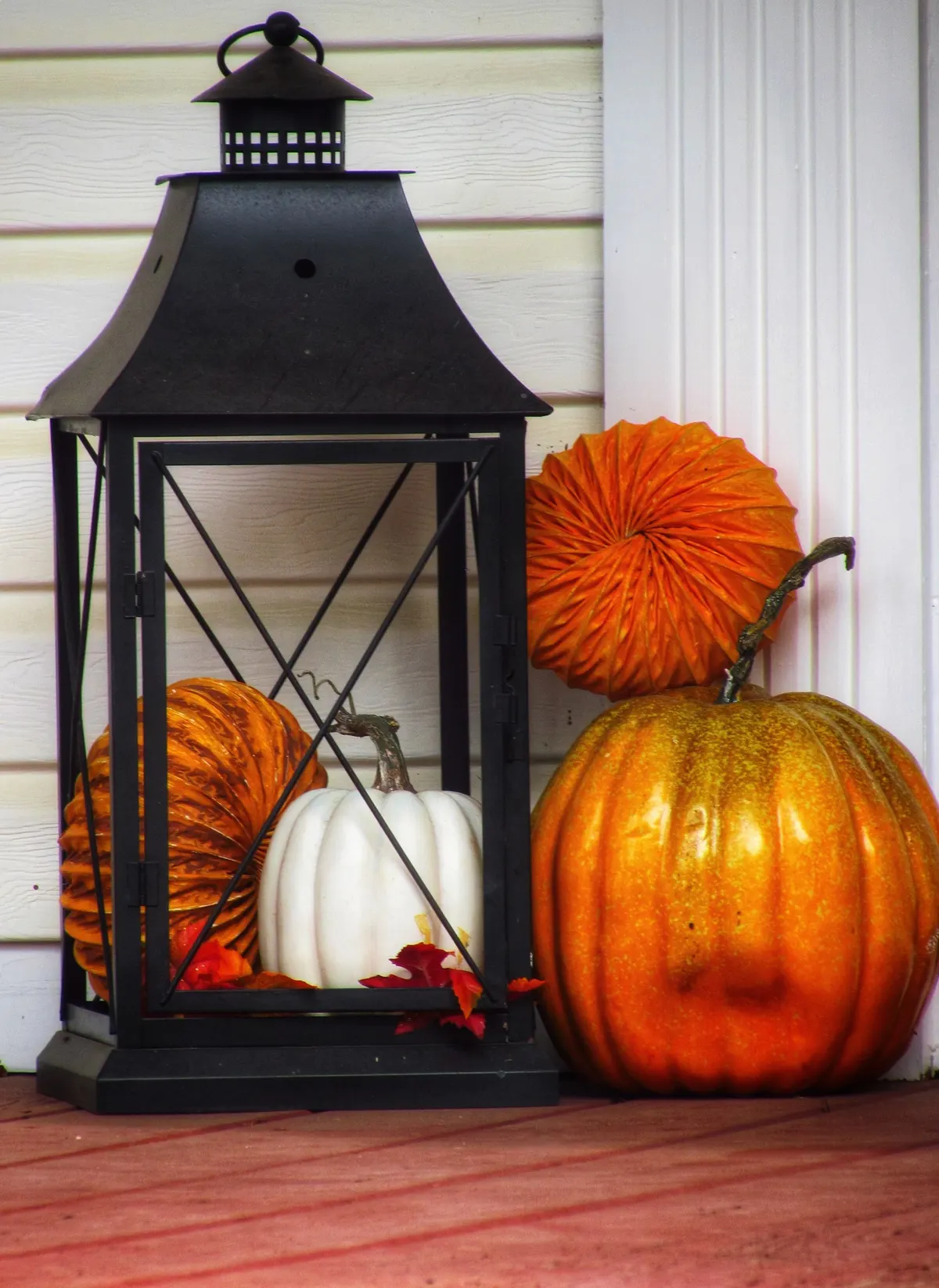 Black metal lantern holding a white pumpkin, surrounded by orange pumpkins on porch decking