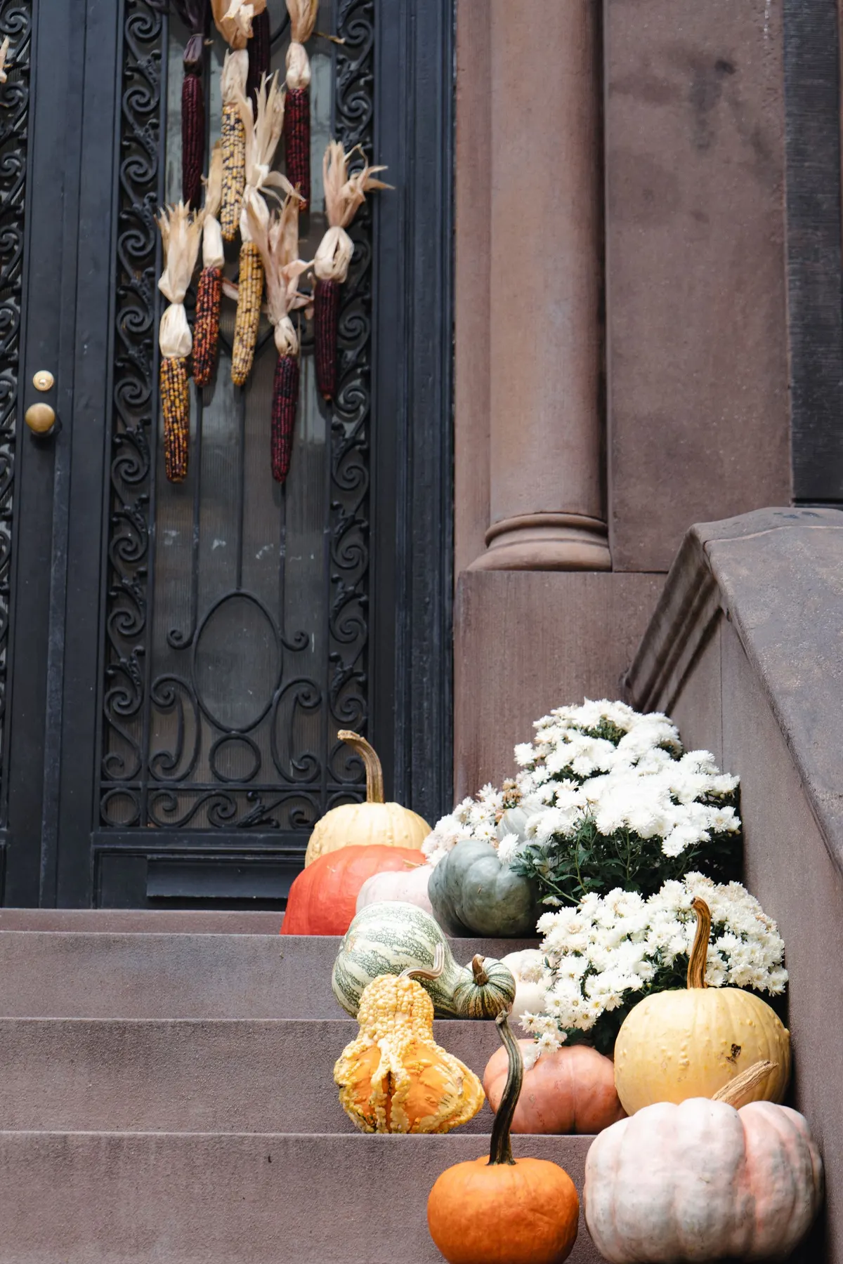 Multicolored pumpkins and white mums lining stone steps below a black door decorated with dried corn