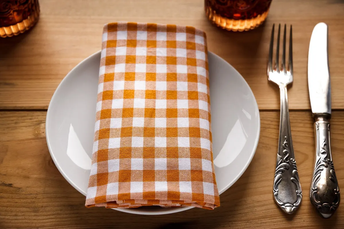 White plate with an orange and white checked napkin and fork beside two amber drinking glasses