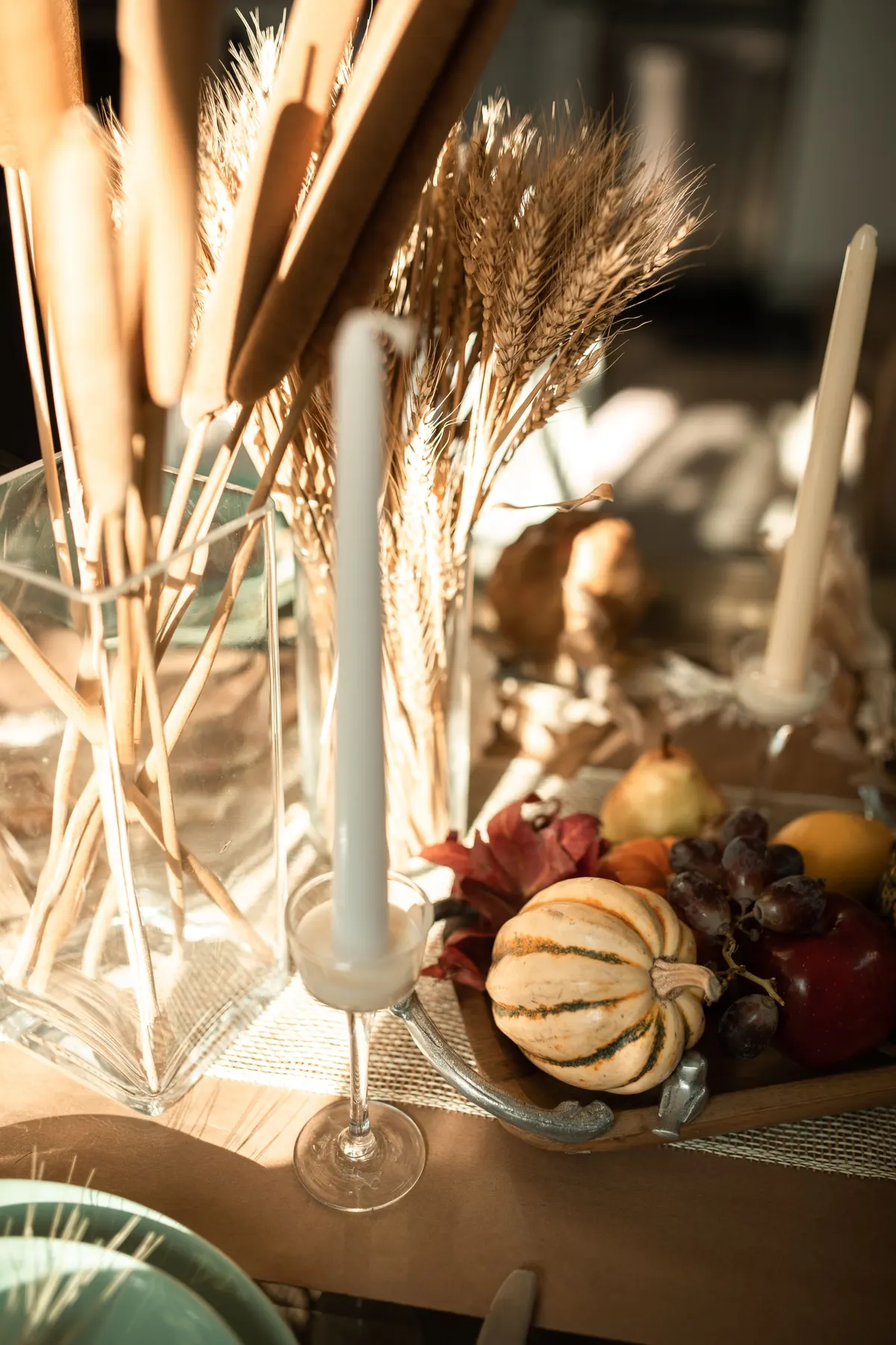 Two glass vases of dried cattails and wheat beside two ivory tapers and a board with a striped pumpkin