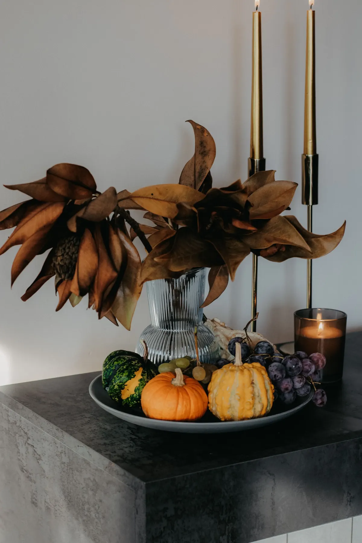 Four gourds, green and purple grapes on a gray platter, a ribbed glass vase and lit tapers