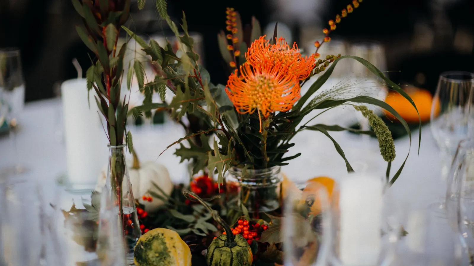 Two glass vases of orange flowers and green foliage with red berries, gourds and white candles on a table