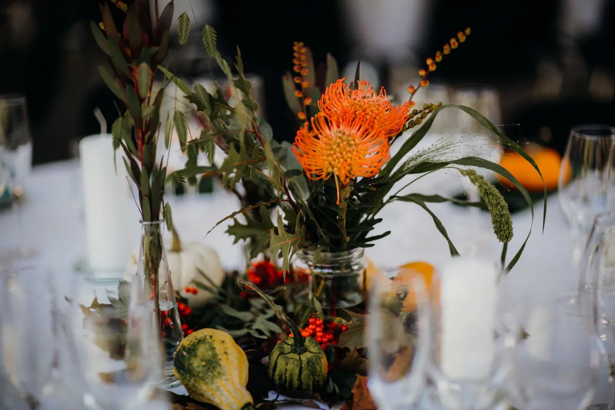 Two glass vases of orange flowers and green foliage with red berries, gourds and white candles on a table