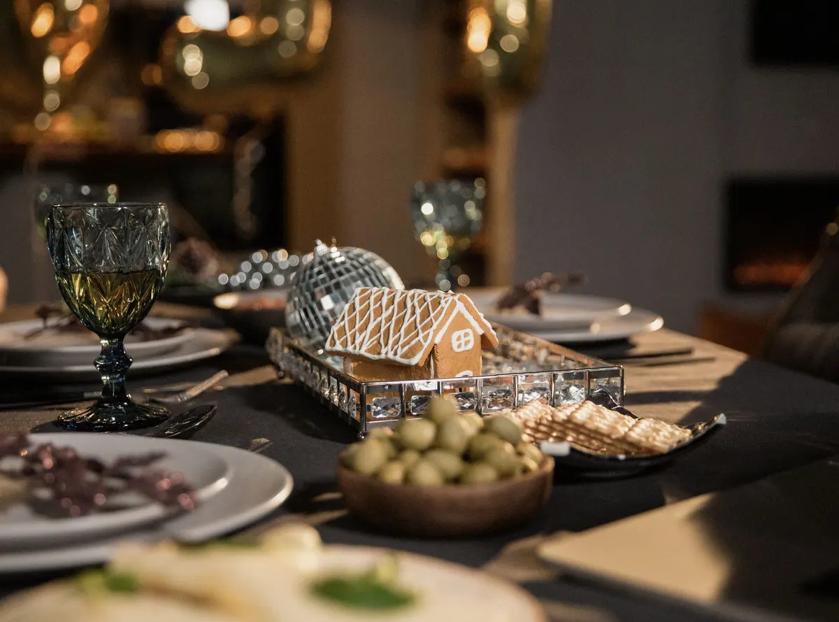 Small gingerbread house with icing lattice roof on a mirrored tray on a dining table with green goblets and a silver ball