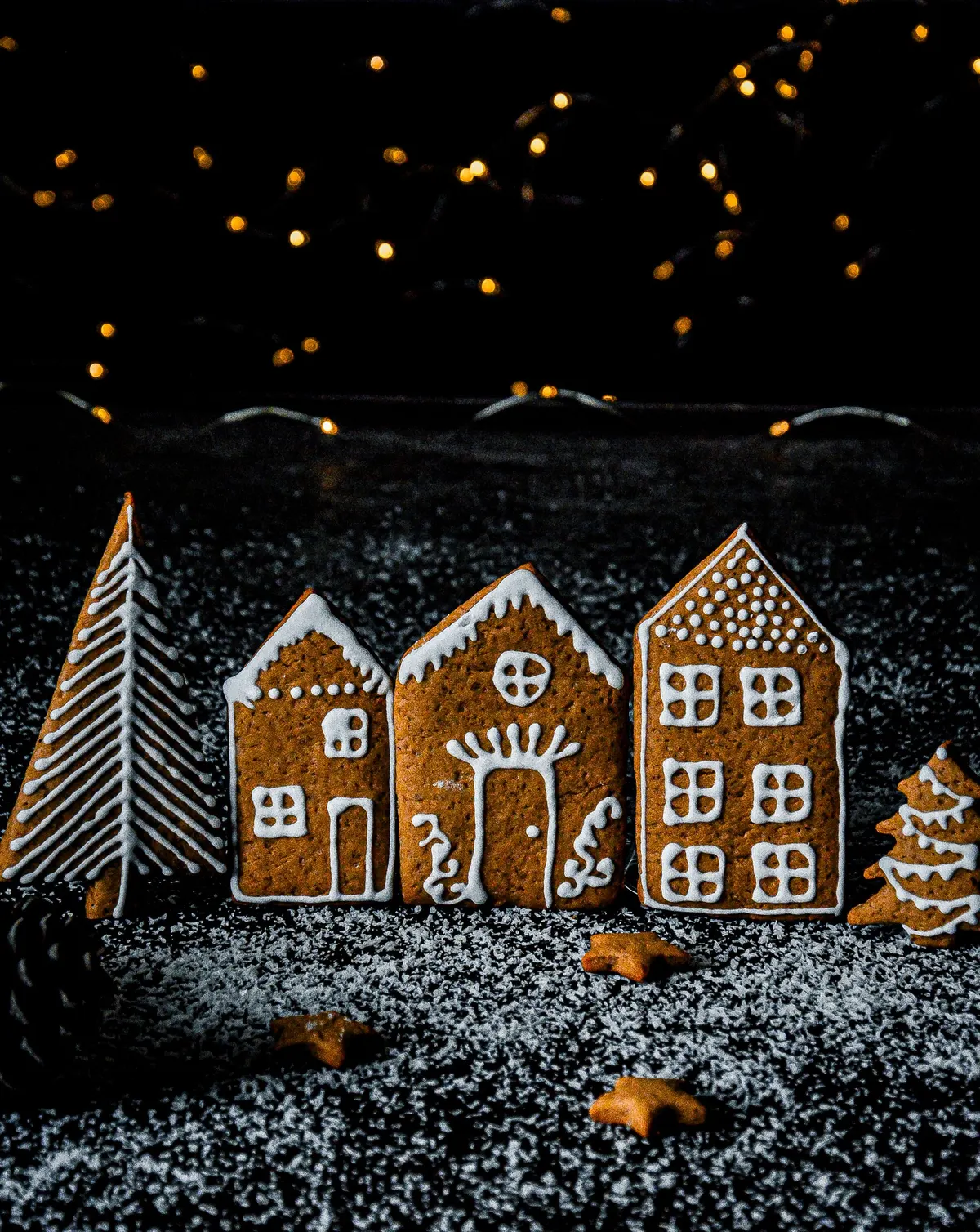 Three flat gingerbread house cookies, two tree cookies and three star cookies with white icing on a dark surface