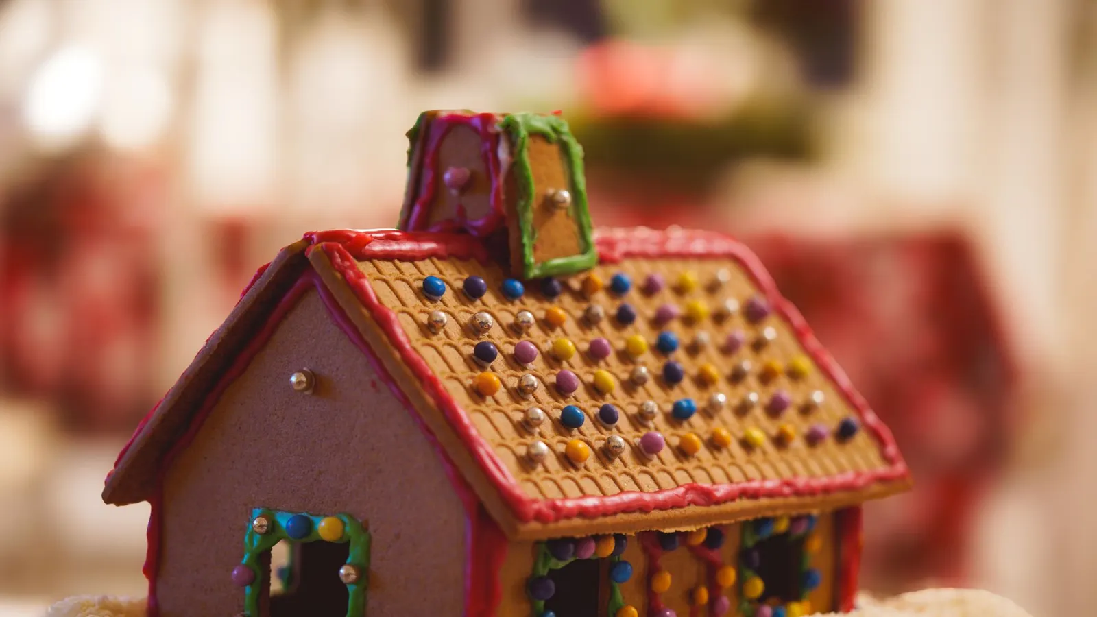 Brown gingerbread house with a chimney, red, green and purple icing and multicolored candy dots, on a tray of white coconut flakes