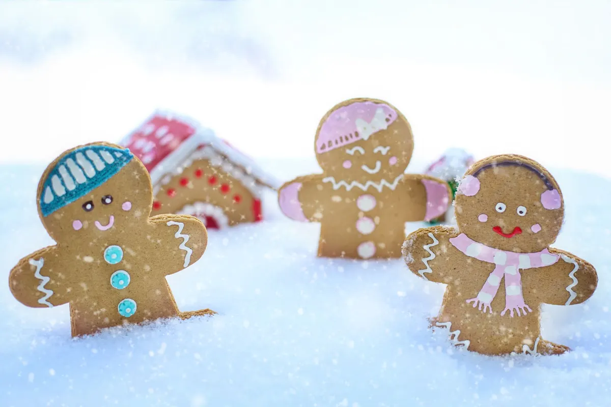 Three gingerbread people with white, pink, turquoise and red icing standing in white snow in front of a gingerbread house