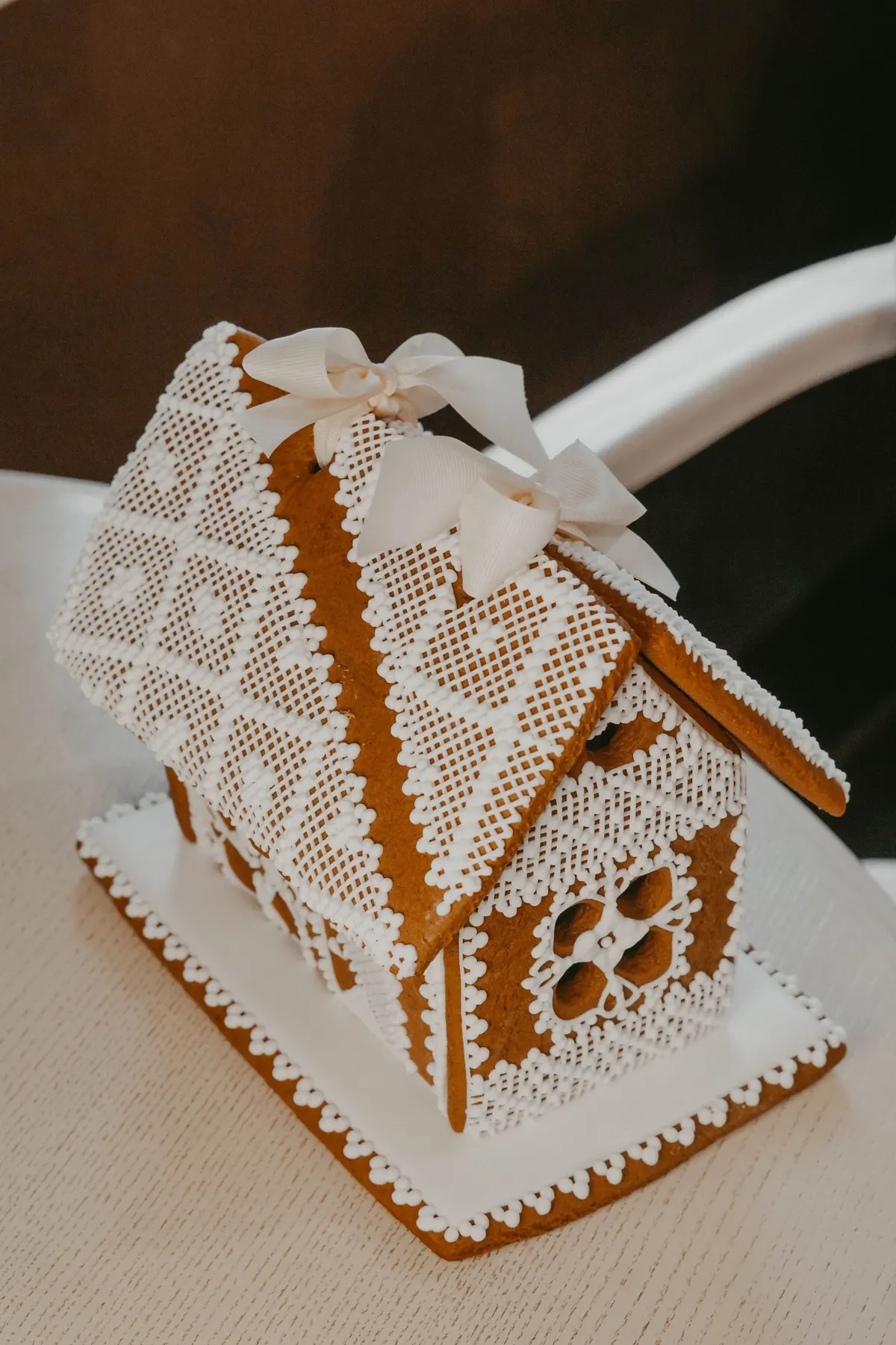 Golden-brown gingerbread house with white icing lattice on the roof, scalloped edging and two ivory ribbon bows