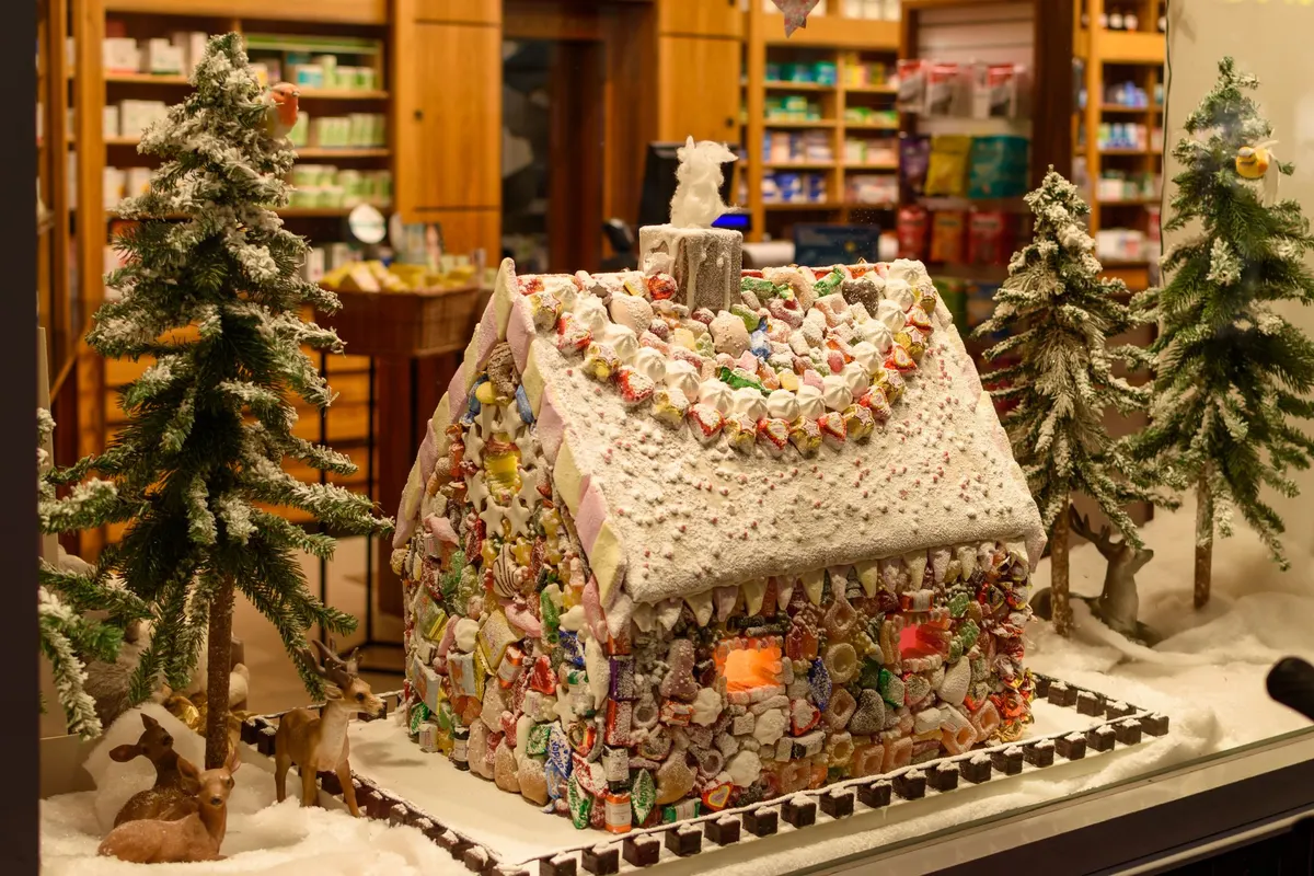 Candy-covered gingerbread house with a snowy white roof and orange-lit window among three snowy fir trees and deer figurines