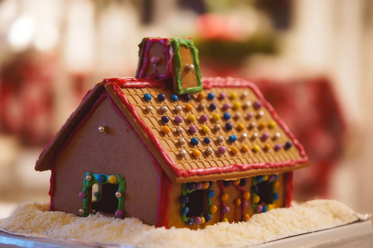 Brown gingerbread house with a chimney, red, green and purple icing and multicolored candy dots, on a tray of white coconut flakes
