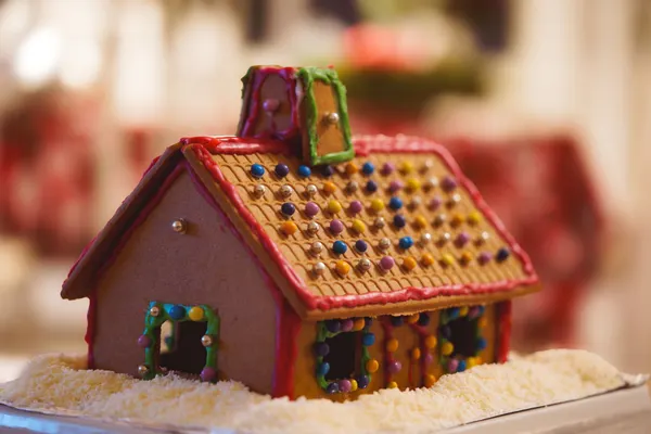 Brown gingerbread house with a chimney, red, green and purple icing and multicolored candy dots, on a tray of white coconut flakes
