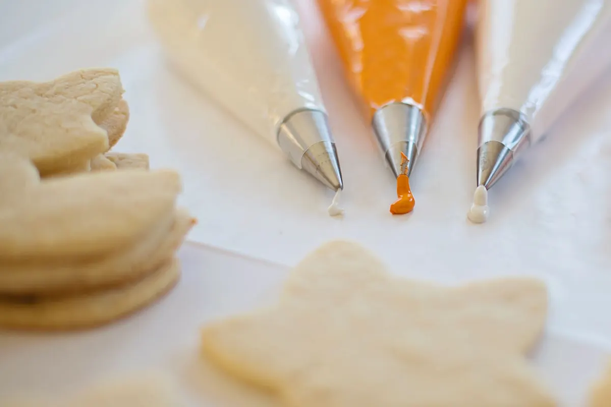 Three piping bags with silver tips holding white and orange icing beside a stack of plain cutout cookies