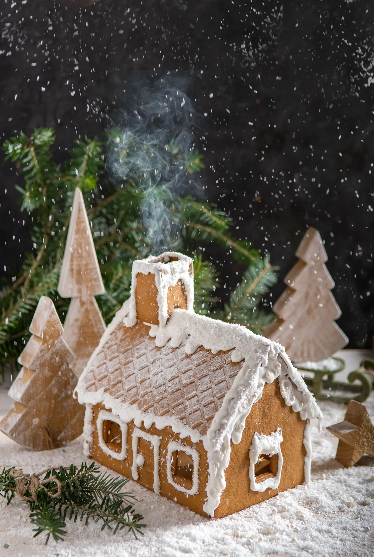 Gingerbread house with white icing, a smoking chimney and three wooden tree cutouts among evergreen branches on a dark surface
