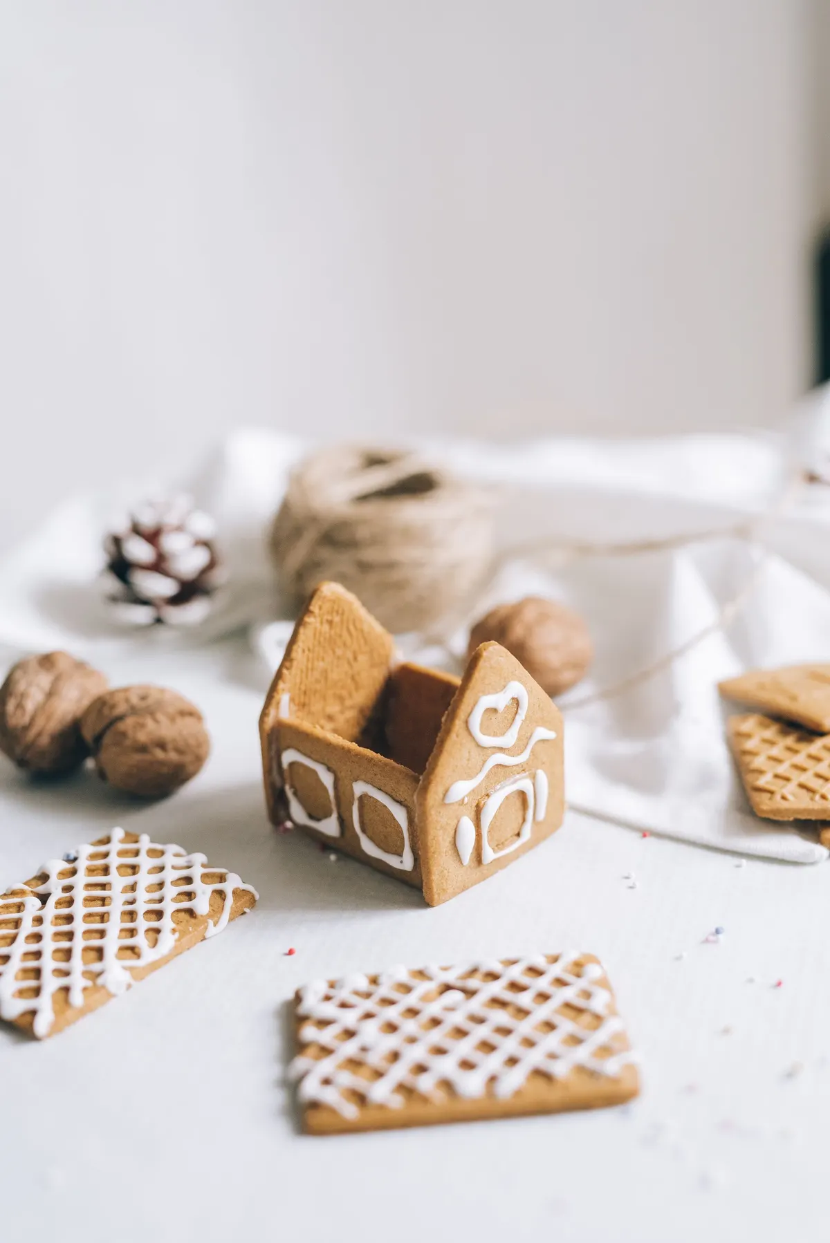 Unfinished gingerbread house beside two iced roof panels, spare biscuit pieces, walnuts, a pinecone and a ball of twine