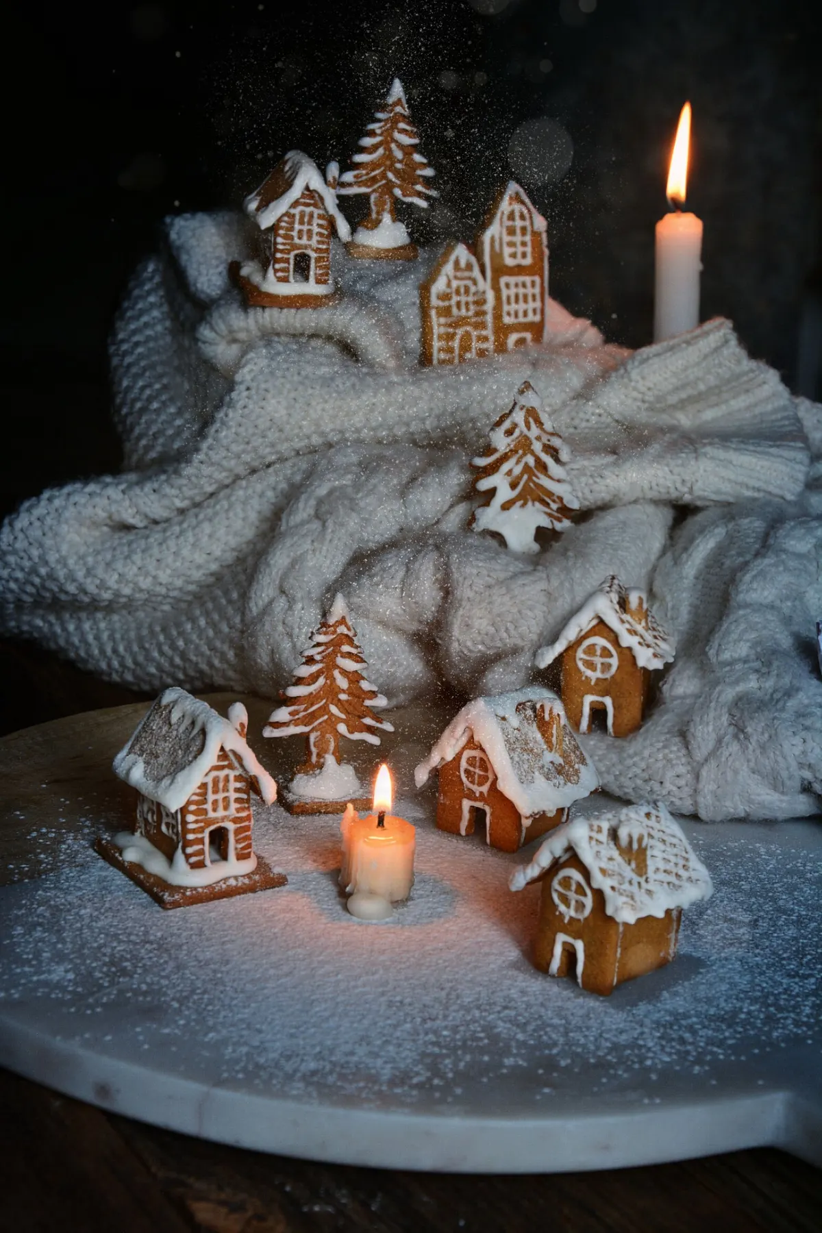 Seven white-iced gingerbread houses and three tree cookies on white knit fabric with two lit white candles