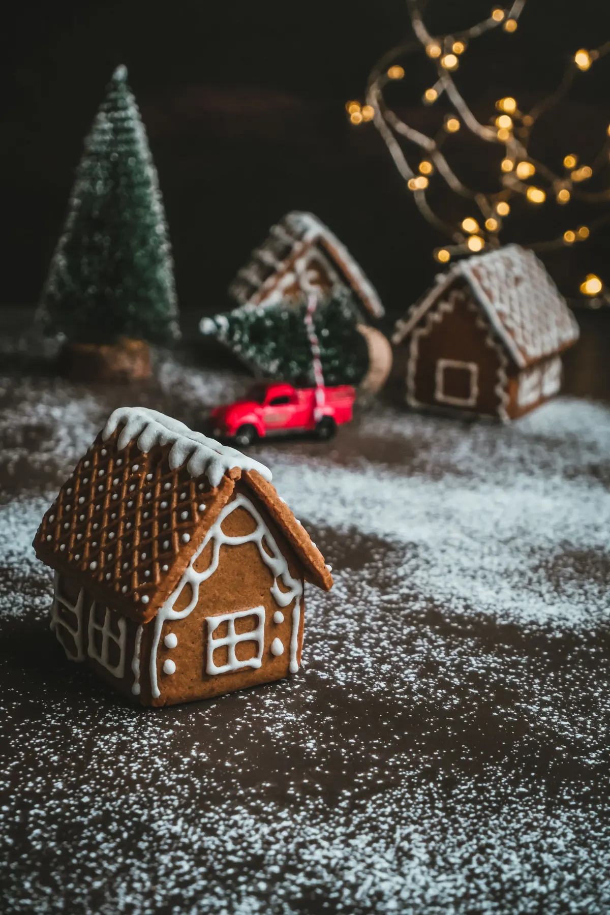 Three gingerbread houses with white icing, two small green trees and a red toy pickup truck on a dusted dark table