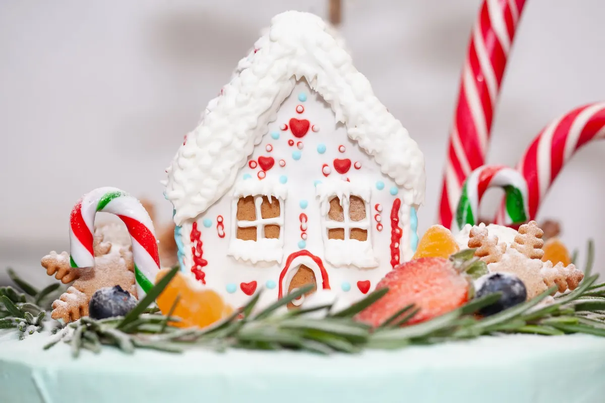 White-iced gingerbread house with red hearts and pale blue dots beside candy canes, two reindeer cookies and green sprigs