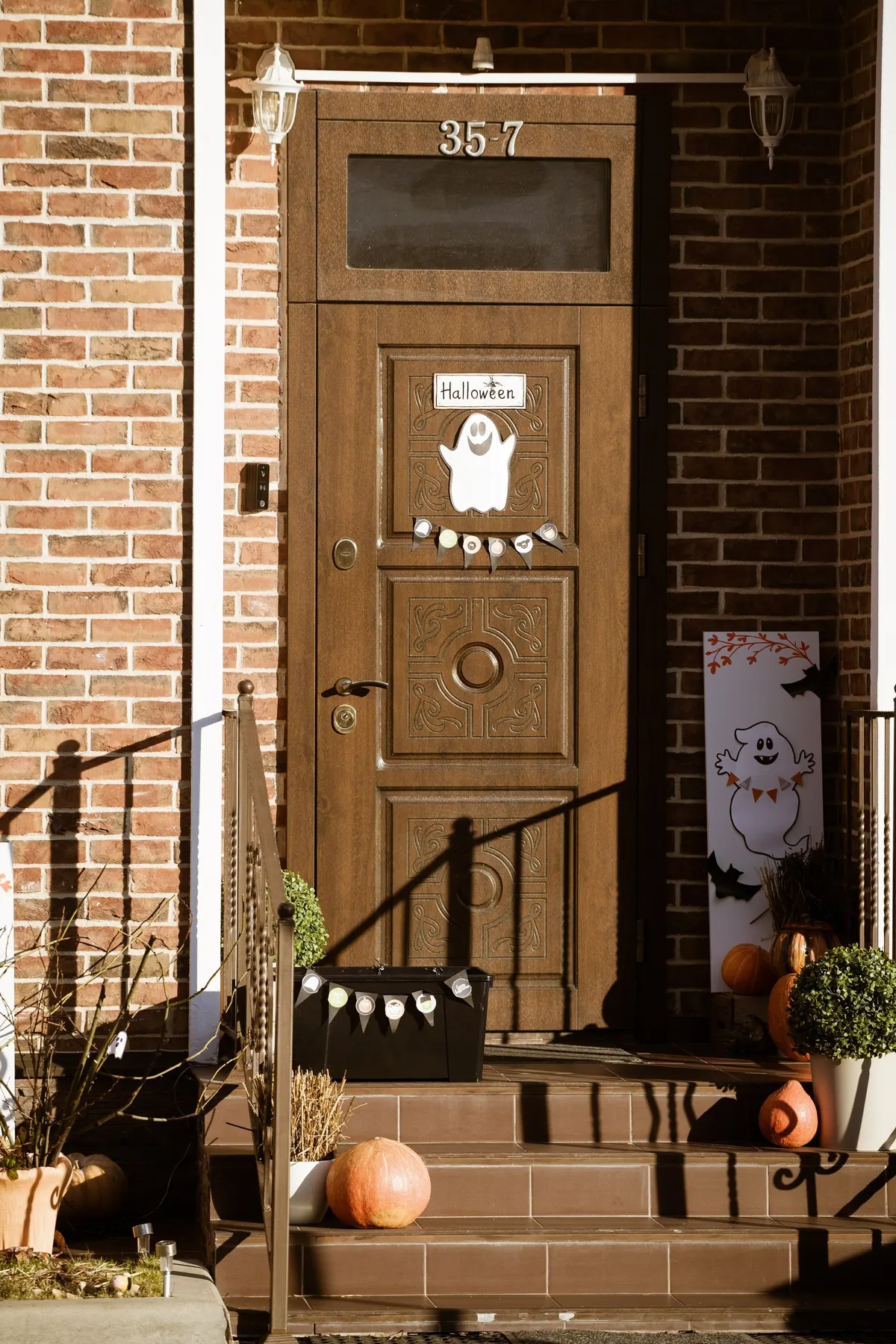 Brown door with a smiling white ghost sign and tiny pennant bunting, a ghost banner and pumpkins on brick steps