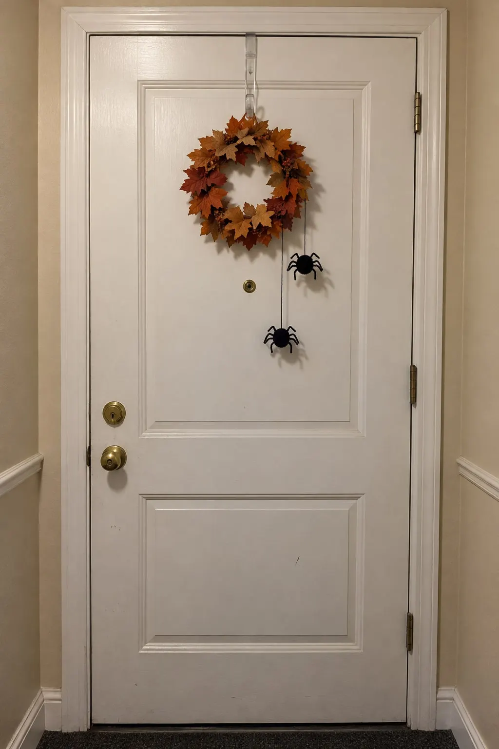 White apartment door with a small leaf wreath and two black pom-pom spiders hanging from a clear over-the-door hook
