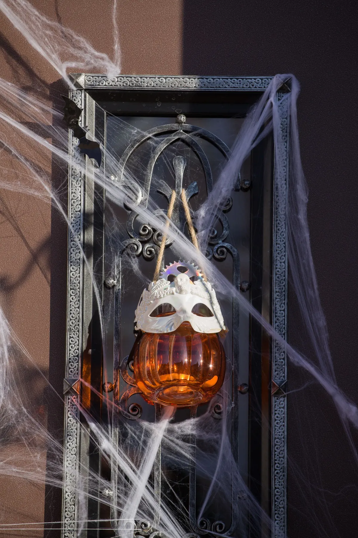 Black wrought-iron door draped in white stretch cobweb, with an amber pumpkin lantern and white mask hanging from rope