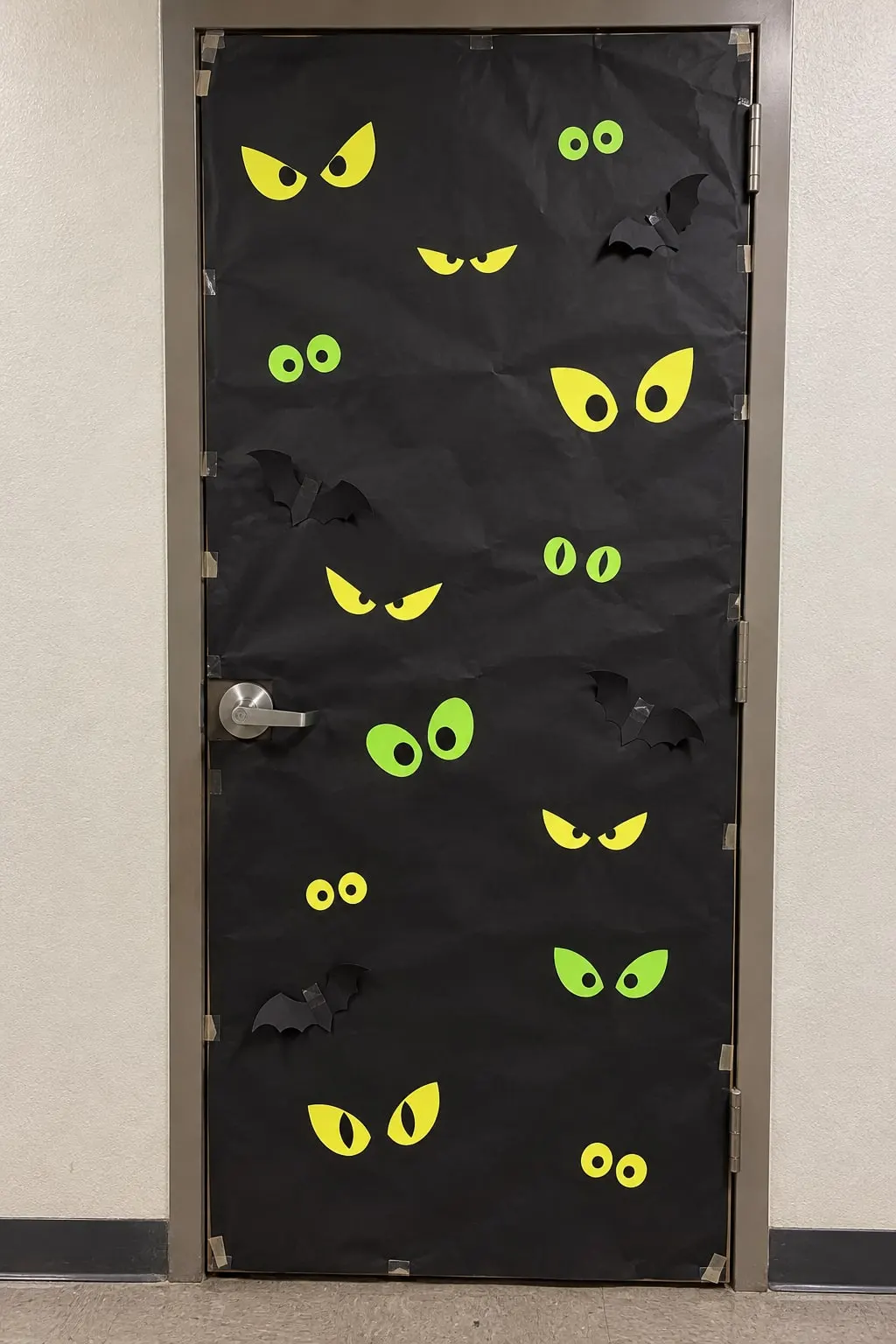 Hallway door covered in black paper with many pairs of yellow and green cut-paper eyes and a few black bats