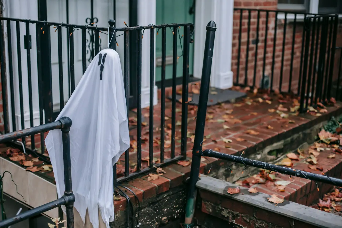 White sheet ghost with black teardrop eyes on a black iron stoop railing with orange mini lights, green door behind