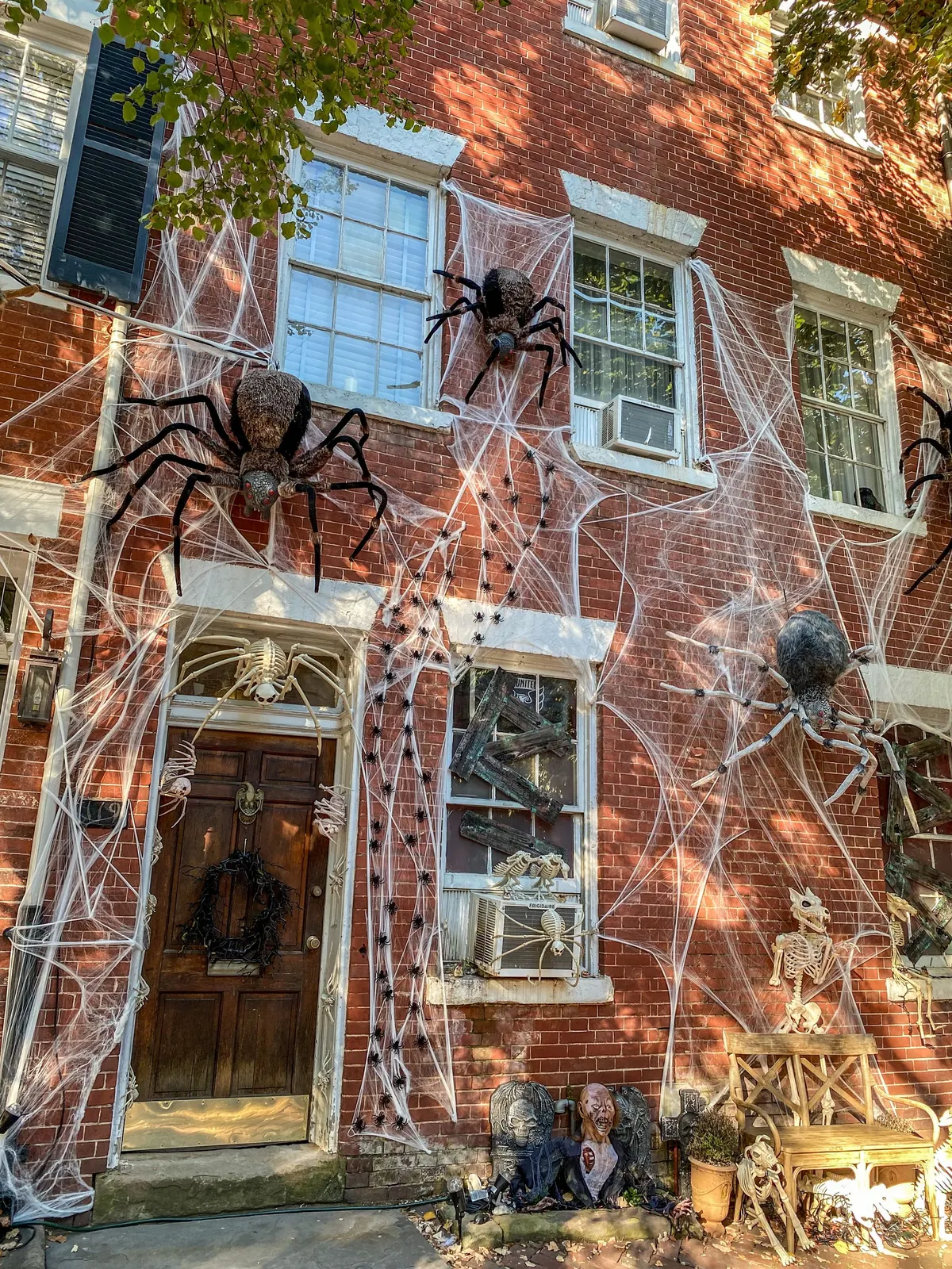 Red brick rowhouse covered in white stretch webbing with four large spiders, a skeleton above a dark door and a twig wreath