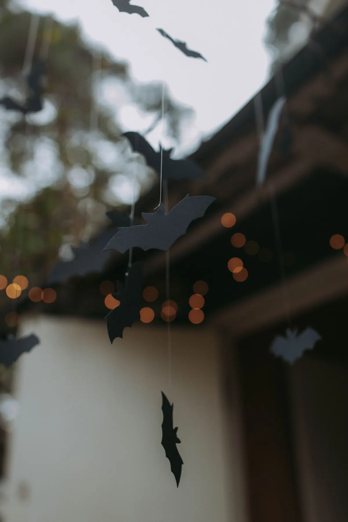 About ten black paper bat cutouts hanging on thin threads in front of blurred foliage and glowing orange lights