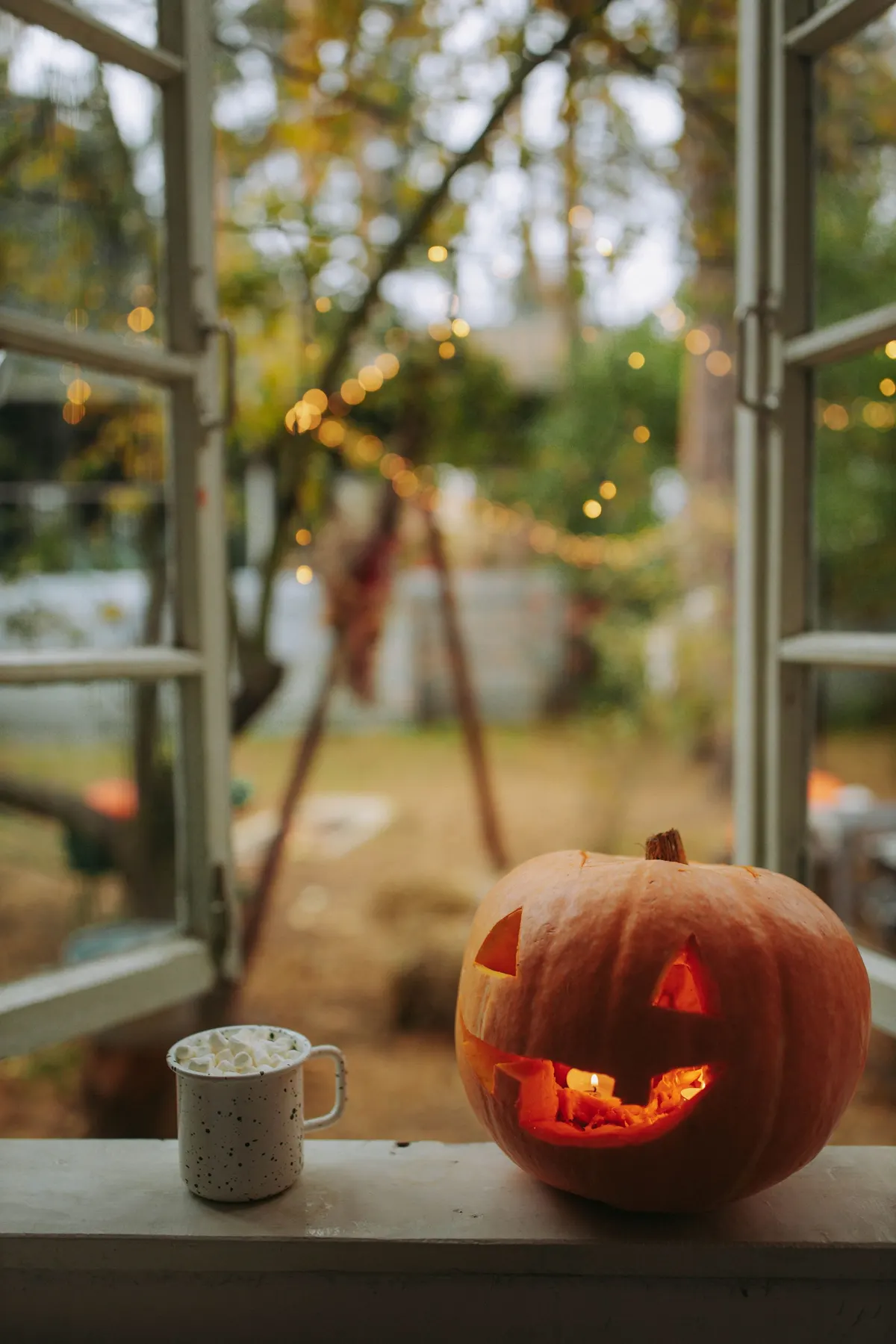 Carved pumpkin with a lit candle and a speckled mug of marshmallows on a window sill