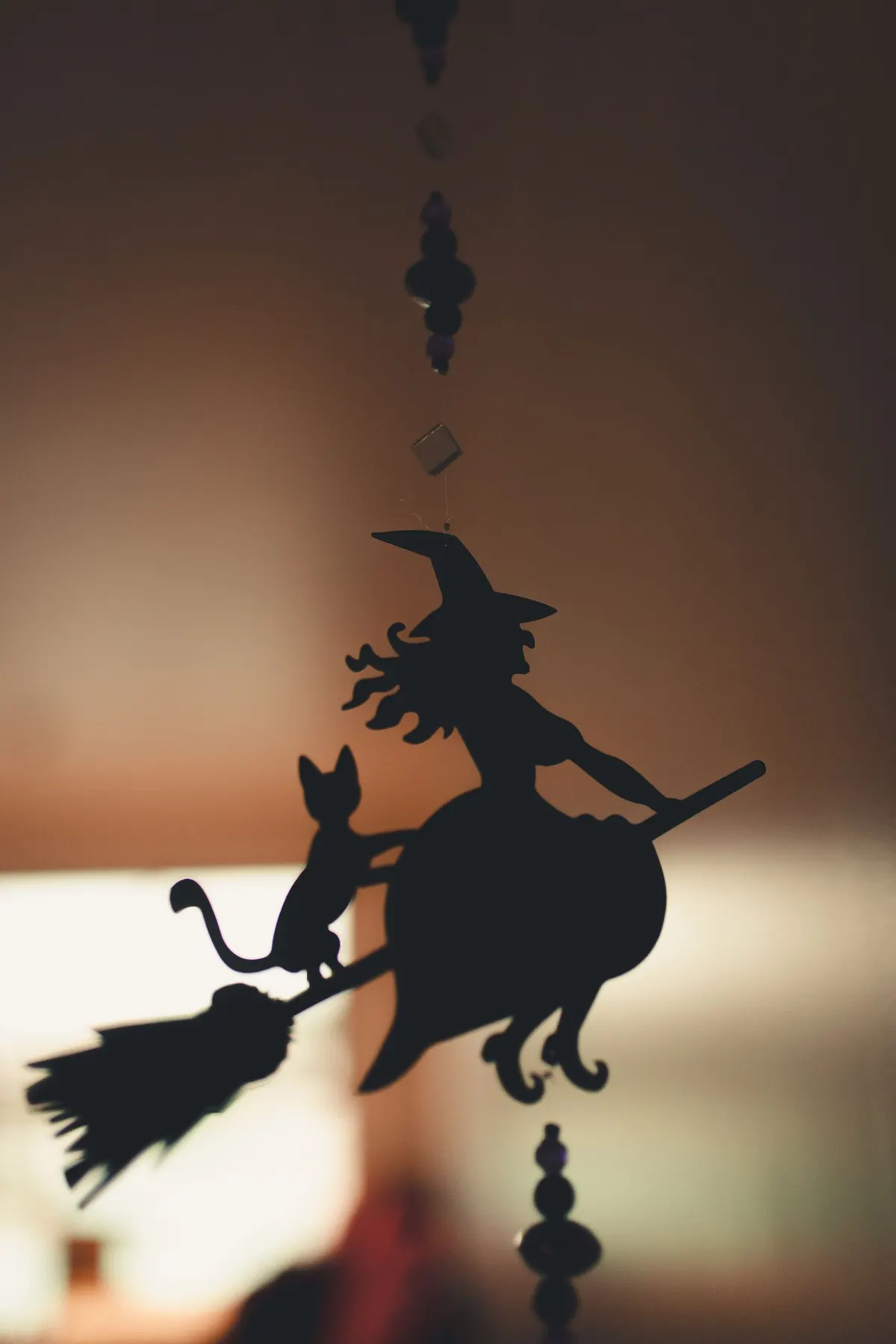 Black silhouette ornament of a pointed-hat witch and a cat riding one broom, hanging on a thread with dark beads