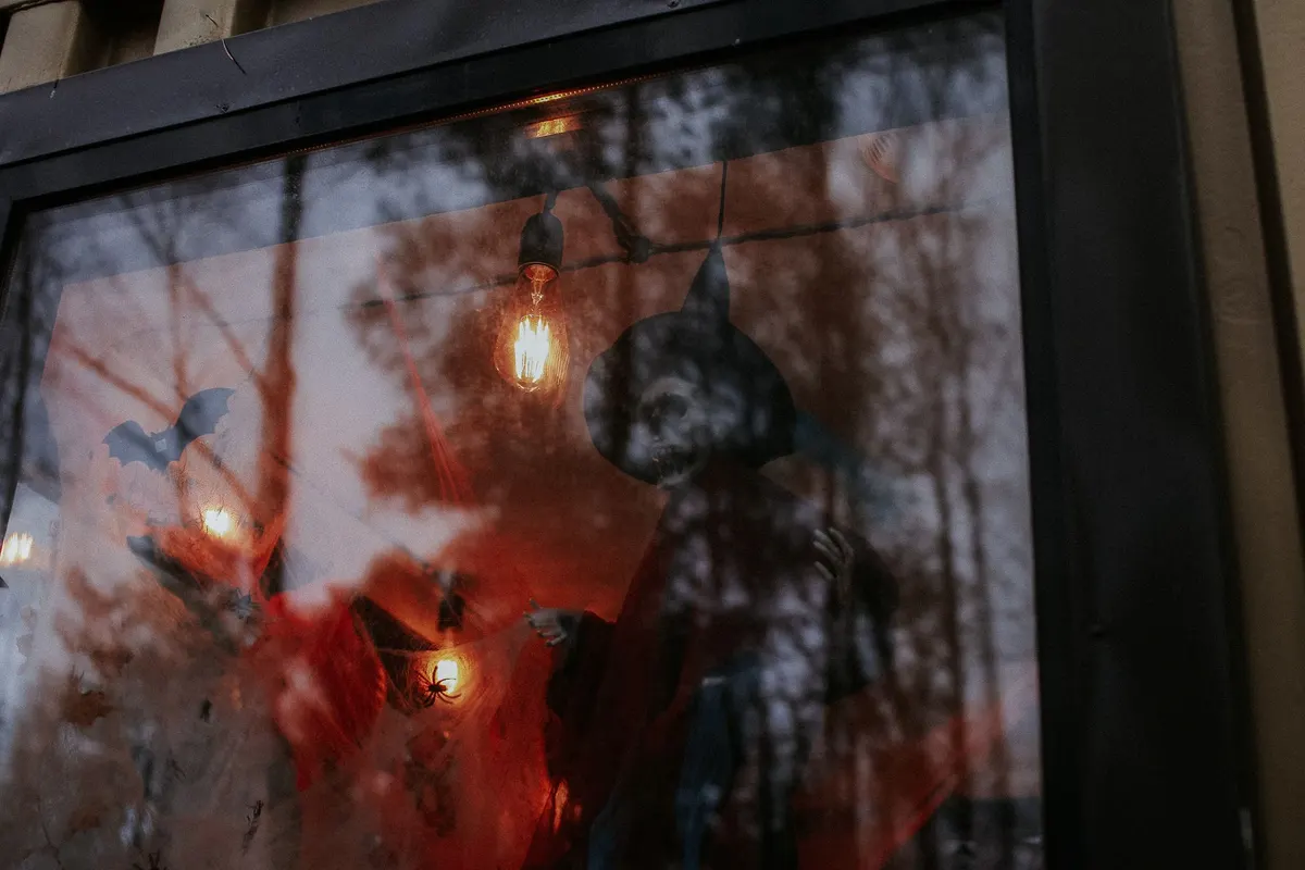 Black-framed window with a skeletal witch in a pointed hat, bat and spider silhouettes, white cobwebs and warm bulbs