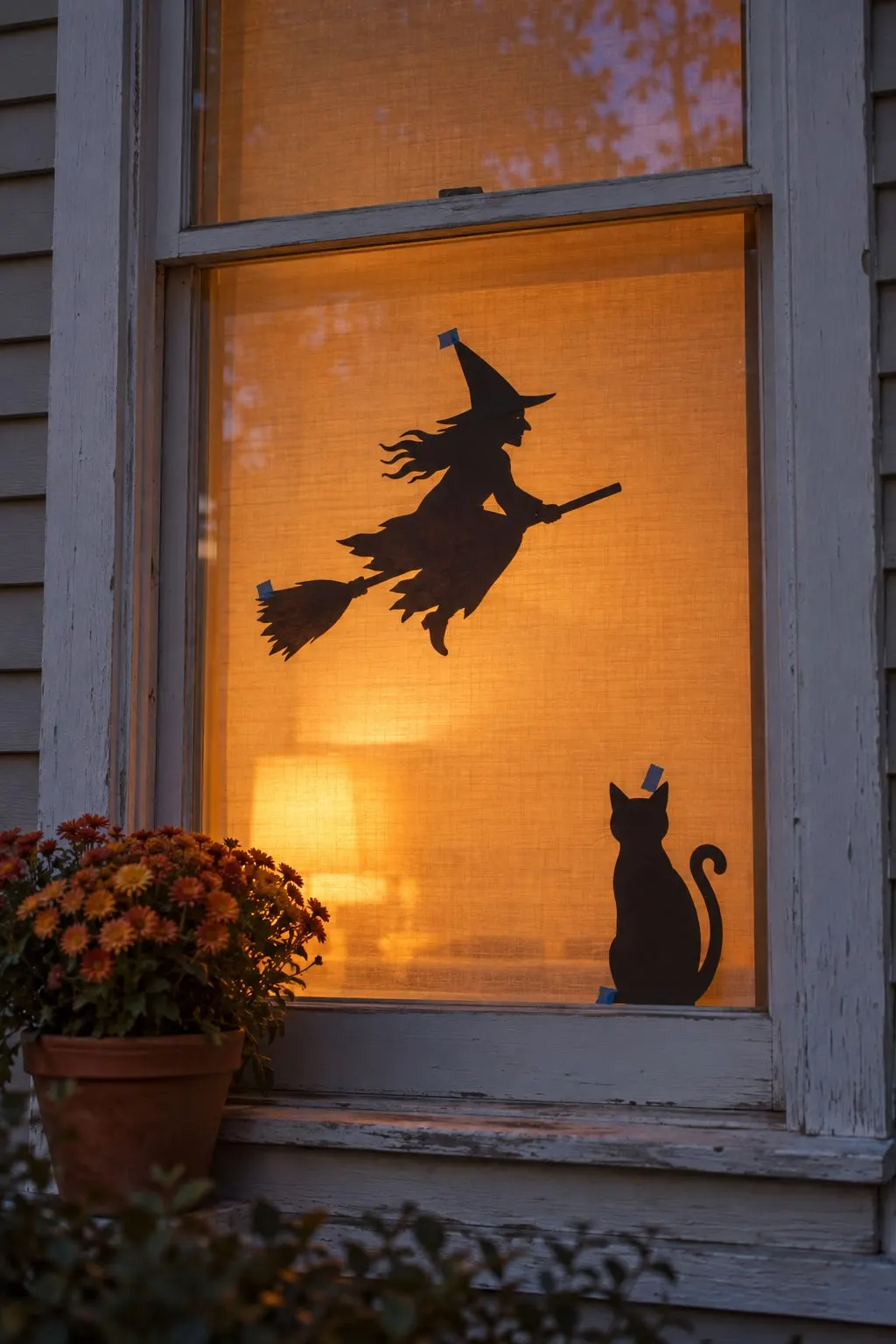 Black paper witch on a broom and a sitting cat taped to window glass, with an orange fabric shade glowing