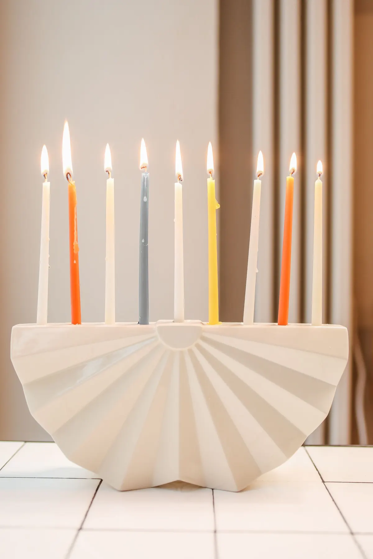 White fluted ceramic menorah with nine lit candles in ivory, orange, yellow and blue on white tiles
