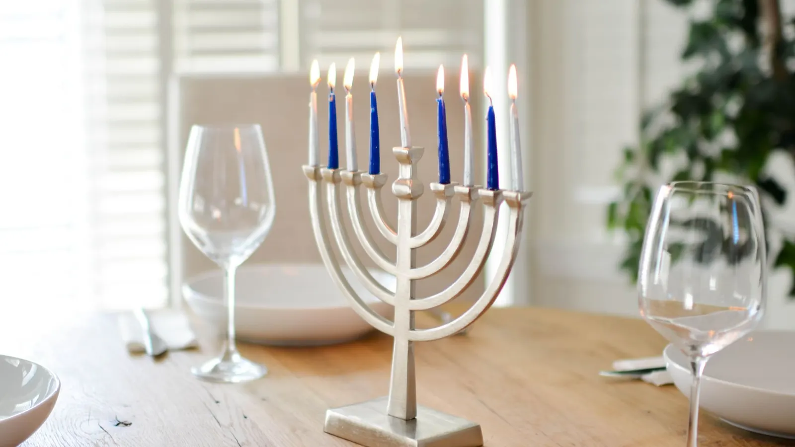 Silver menorah with four blue and five white lit candles on a dinner table with wine glasses