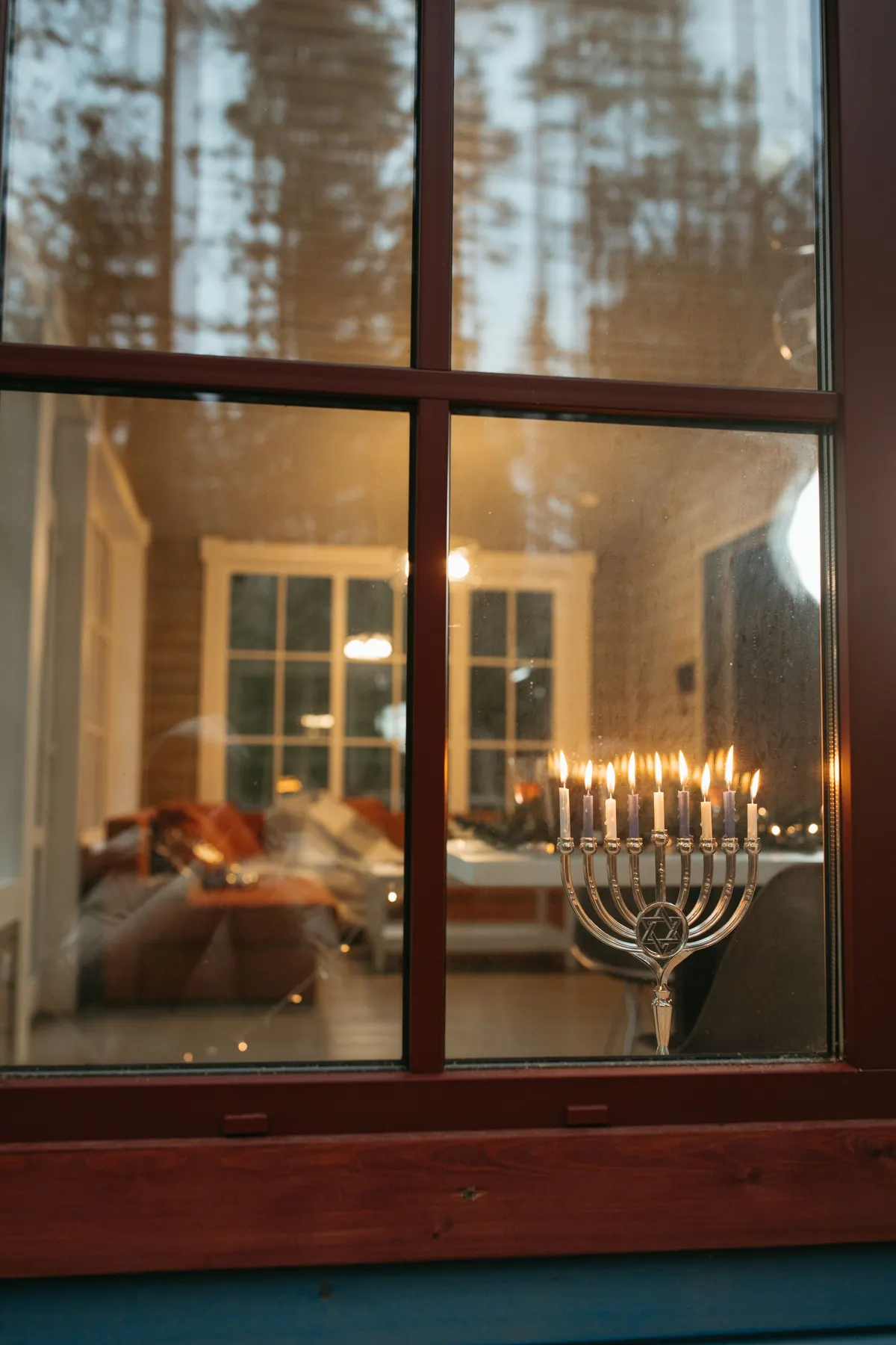Silver menorah with a Star of David center and nine lit candles seen through a wooden window frame