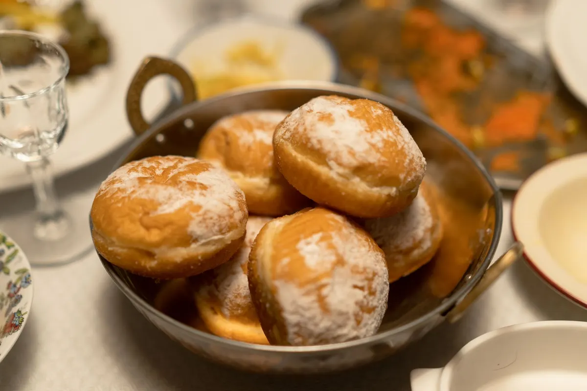 Six powdered sugar doughnuts piled in a round two-handled metal pan beside a stemmed glass