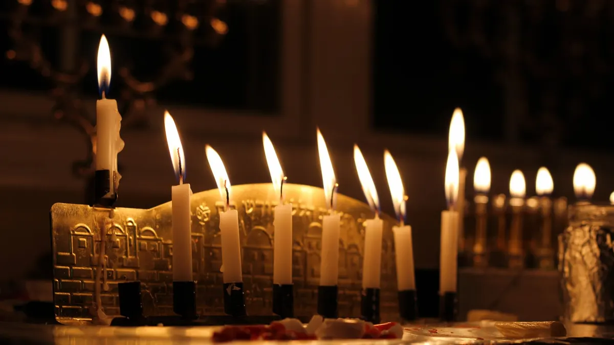Gold menorah with eight lit cream candles in front of several more glowing menorahs on a shared surface